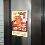 Clear acrylic wall-mounted poster frame displaying a bakery menu with croissants and pricing, designed for clean and professional in-store advertising